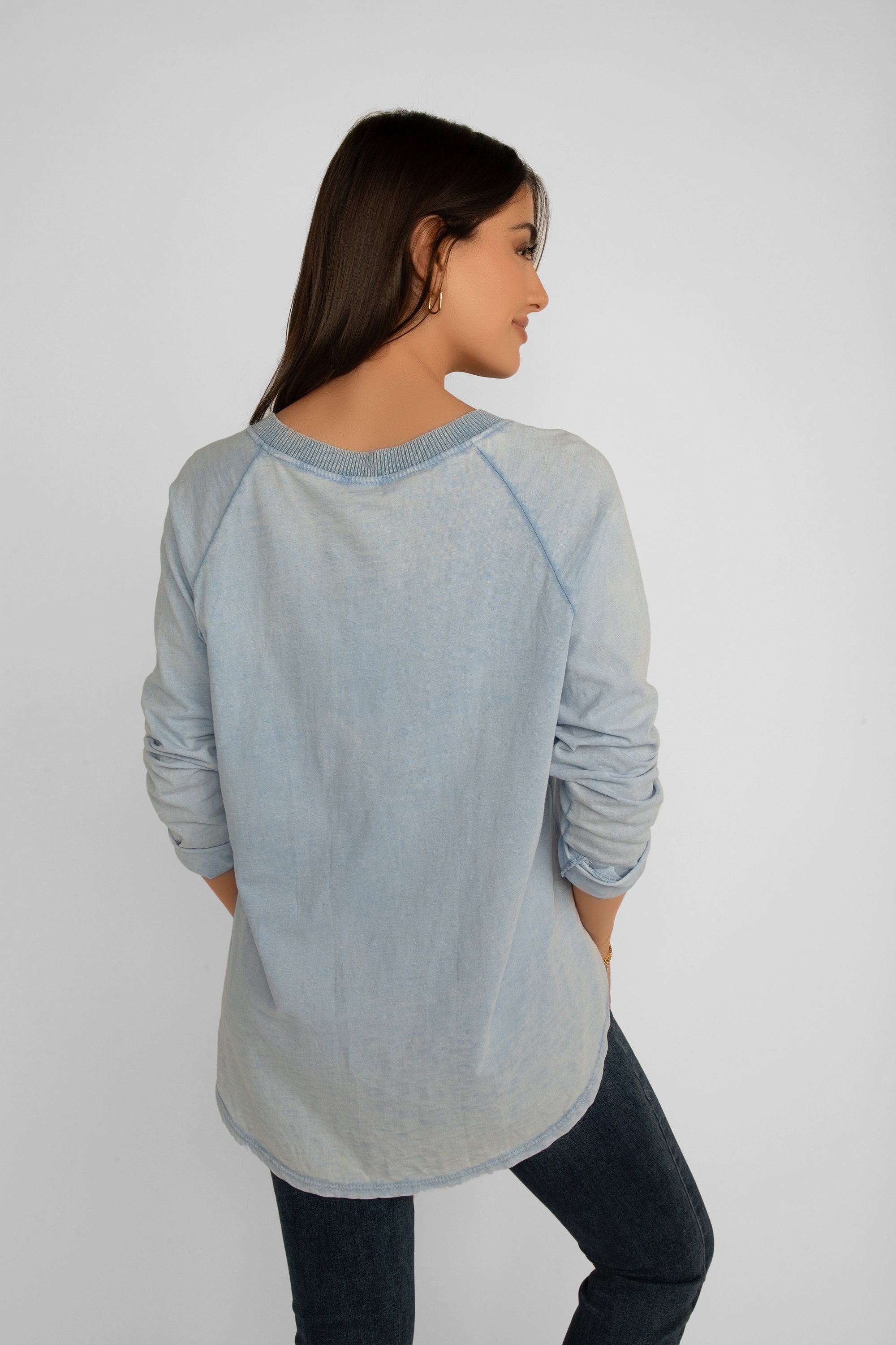 Back view of Elissia (JY3734) Women's Long Raglan Sleeve Garment Dye V-Neck Top with Button Patch Detail on left hip, and a high low hem in Baby Blue